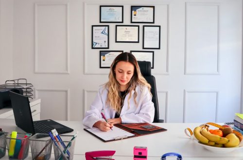Nutritionist sits at a modern office desk writing, with fruits and diplomas displayed, emphasizing health and professionalism.
