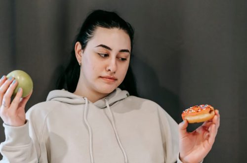 Contemplative plump female in hoodie choosing between ripe green apple and delicious donut in studio