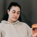 Contemplative plump female in hoodie choosing between ripe green apple and delicious donut in studio